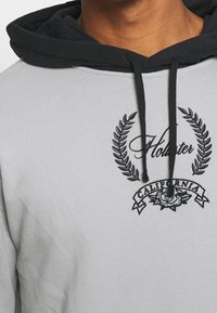 Sudadera gris claro con una capucha negra, que presenta el texto bordado en negro "Hollister" y un diseño decorativo de laurel con el texto "California" abajo.