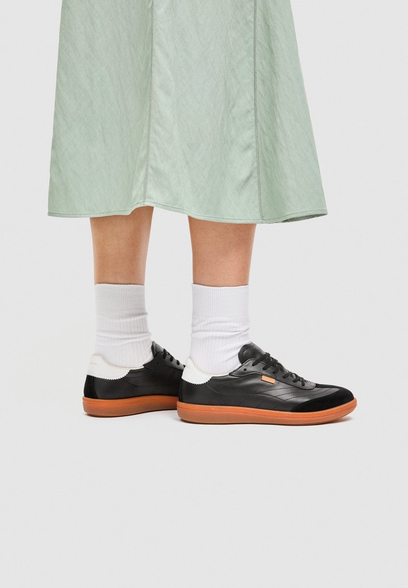 Ben iført en lys grøn nederdel, hvide sokker og sorte lave sneakers med brune såler mod en enkel baggrund.