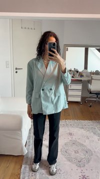 Göndör sötét hajú személy világoskék zakót, fekete nadrágot és sneakers-t viselve tükörben készít önarcképet egy világos szobában, ahol bútorok is találhatóak.