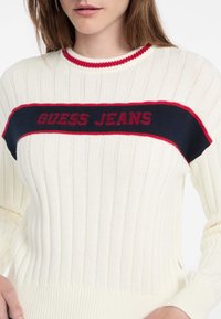 Pulover crem cu dungi ribbed, având o bandă roșie și bleumarin pe piept, cu textul "GUESS JEANS". Mâneci lungi și design ajustat.