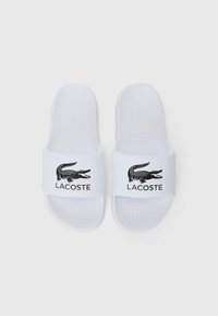 Vita slide-sandaler i vitt med en texturerad fotbädd och ett brett rem med en svart krokodil-logotyp och "LACOSTE" tryckt under.