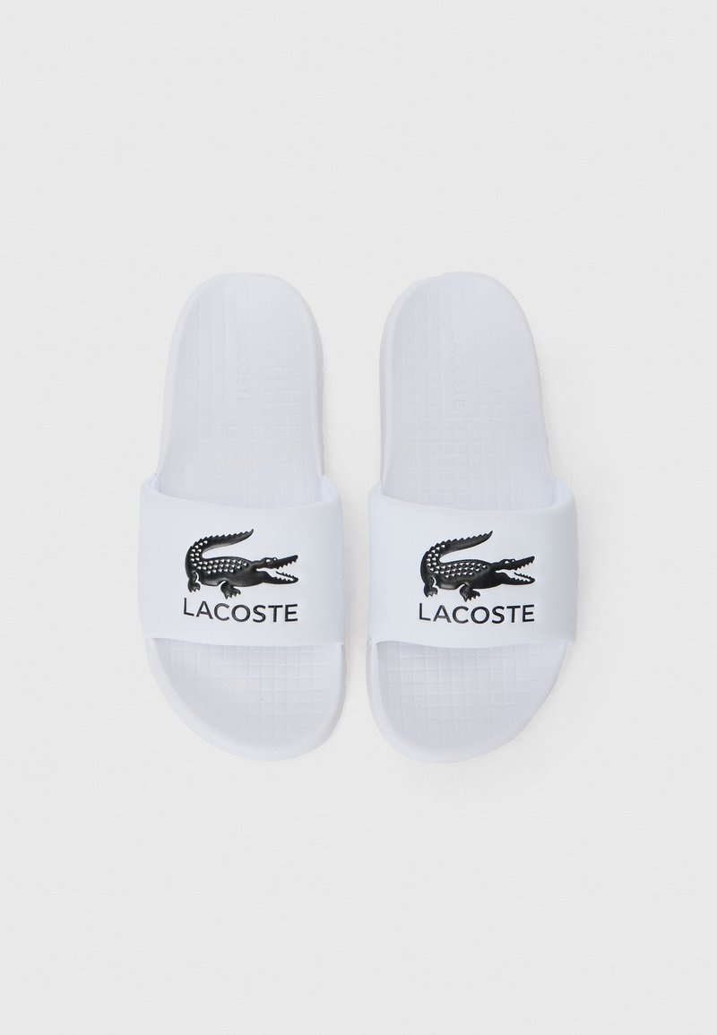 Weiße Rutschsandalen mit einer strukturierten Innensohle und einem breiten Riemen, der ein schwarzes Krokodil-Logo und "LACOSTE" darunter aufgedruckt hat.
