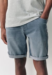 Hellblaue Jeansshorts mit aufgerollten Säumen, lockerem Schnitt und Fünf-Taschen-Design. Kombination mit einem gestreiften T-Shirt mit feinen schwarzen Linien.