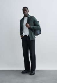 Pantalones vaqueros negros, camiseta blanca, camisa verde abotonada y bolso de cuero negro con acabado texturizado. El modelo está de pie frente a un fondo neutro.