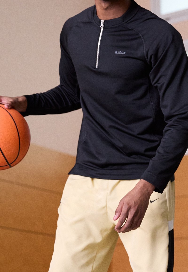 Černé sportovní tričko s dlouhým rukávem a čtvrtovým zipem vpředu, s malým logem, v kombinaci se světlými žlutými kalhotami s bočním logem. Drží basquetbal.