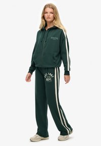 Groene zip-up hoodie met witte sportieve letters, gecombineerd met bijpassende donkergroene broek met witte zijstrepen en logo-detail.