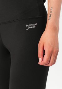 Juodos tamprės, pagamintos iš elastingos medžiagos, su sidabriniu Tommy Jeans logotipu apatinėje pusėje. Lygus paviršius ir prigludusi forma.