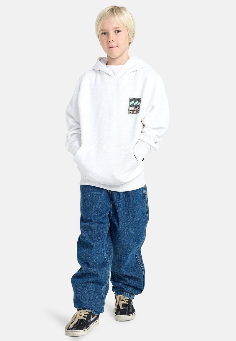 Witte hoodie met een voorzak en een blauw-zwart grafisch patch, gecombineerd met losse blauwe spijkerbroek en zwarte sneakers.