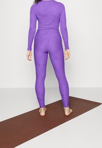 Purple athletic set featuring long sleeves and fitted leggings with textured patterns, displayed on a yoga mat. Smooth, stretchy fabric.