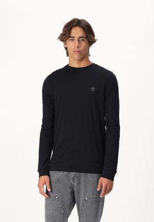 Svart langermet t-skjorte laget av myk stoff, med ribbet rund hals og et lite emblem på brystet, kombinert med grå bukser.