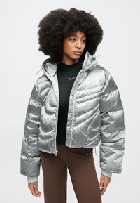 Hopeanvärinen, pehmustettu takki, jossa on lyhyt malli, huppu ja etuvetoketju. Takissa on tikattu pinta ja joustavat hihansuut, ja se on puettu mustan paidan päälle.