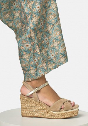 Pied bronzé avec une sandale compensée en daim beige, plateforme en jute tressé et bride à la cheville, porté avec un pantalon à motif floral turquoise.