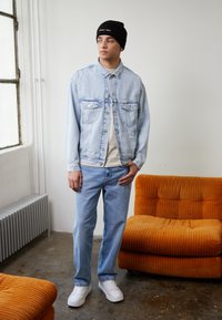 Giacca di jeans blu chiaro con due tasche sul petto, abbinata a jeans blu chiaro e sneakers bianche. Berretto nero con accento di testo. Divano in corduroy arancione.