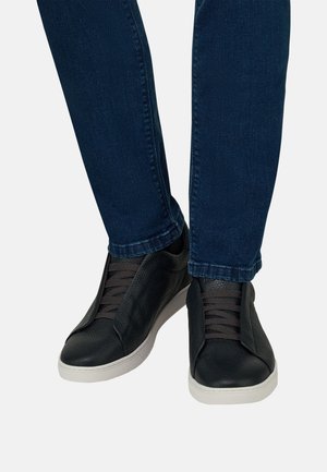 Persona che indossa jeans blu scuro e sneakers in pelle nera testurizzata con suole bianche su uno sfondo chiaro e uniforme.