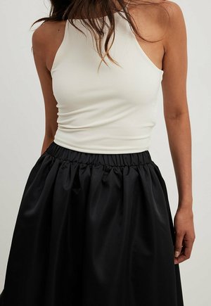 Woman wearing a sleeveless white halter top tucked into a high-waisted black gathered skirt, standing against a neutral background.