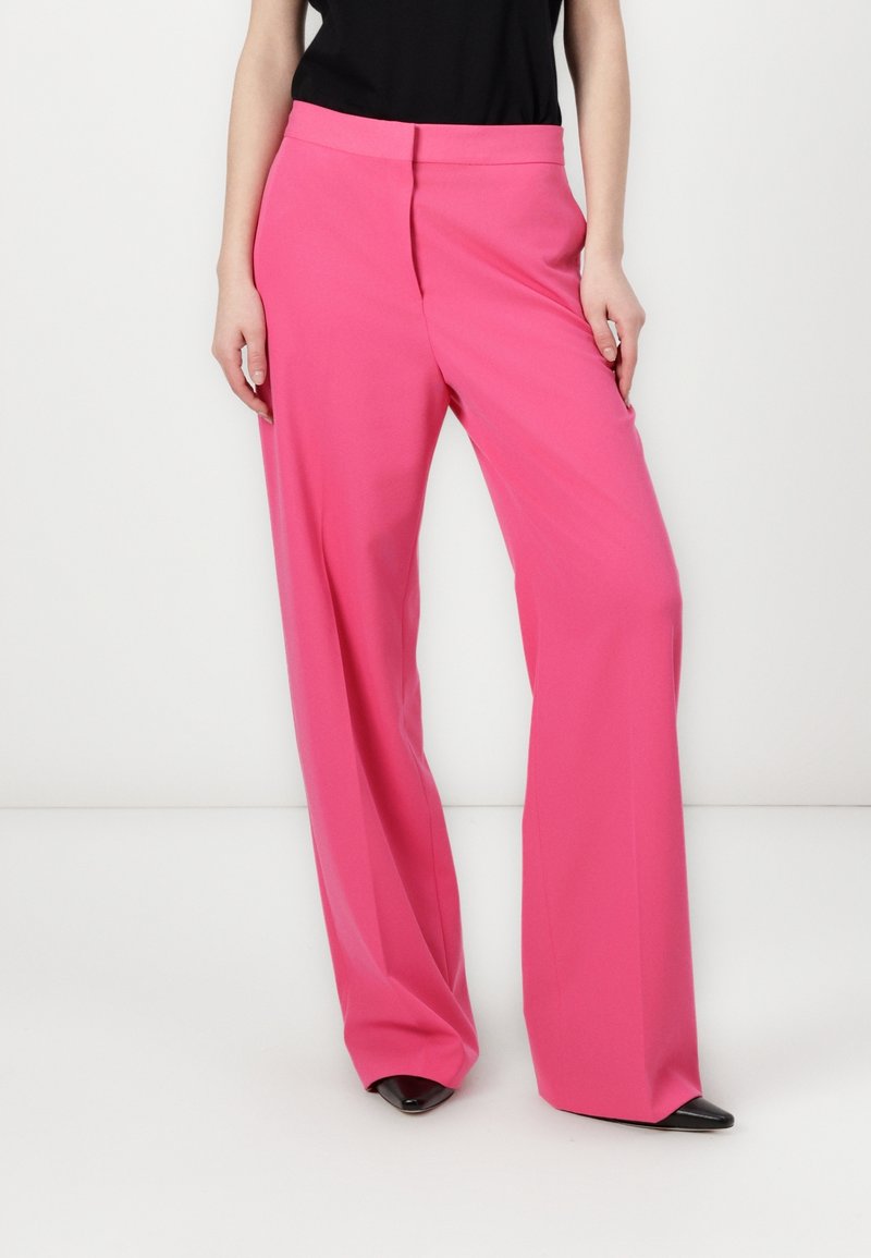 Femme portant un pantalon taille haute, large et rose vif, avec une chaussure noire à bout pointu visible sous l'ourlet.