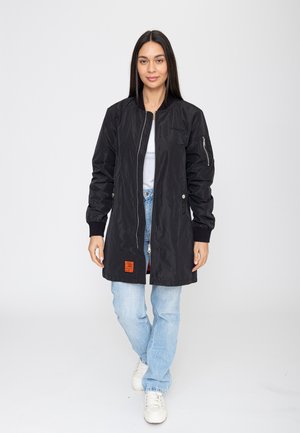 Frau mit langen dunklen Haaren, die eine schwarze Bomberjacke mit Reißverschluss, ein weißes Hemd, hellblaue Jeans und weiße Sneakers trägt, steht vor einem weißen Hintergrund.