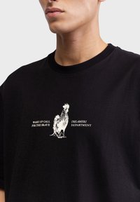 Zwart katoenen T-shirt met een witte afbeelding van een haan en de tekst "WAKE UP CALL FOR THE BRAVE" en daaronder "DREAMERS DEPARTMENT".