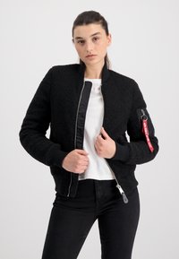 Zwarte bomberjack met een donzige buitenkant, ritssluiting, geribbelde manchetten en een contrasterend satijnen paneel. Heeft een rode label op de mouwzak.