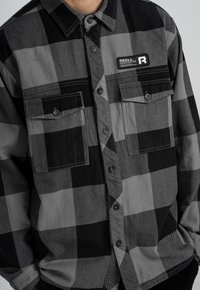 Schwarz-graues karierter Flanellhemd mit zwei Brusttaschen, Knopfverschluss und einem kleinen Logo-Patch auf der linken Seite. Strukturierter Stoff.