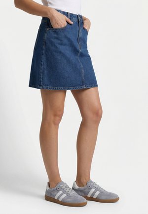 Kvinde iført en blå denimnederdel, hvid top og grå sneakers med hvide striber, stående med hænderne i nederdelslommerne.