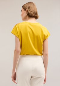 Geel kortemouwen shirt met een relaxte pasvorm en decoratieve accenten op de schouders. Draag met crème kleurige broeken. Glad stoffen textuur.