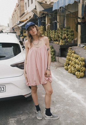 Różowa sukienka bez rękawów o luźnym kroju, zestawiona z czarno-białymi trampkami w kratkę, stoi obok białego samochodu na tle targu.