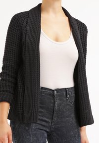 Schwarzer gestrickter Cardigan aus strukturiertem Stoff, mit offenem Vorderteil und langen Ärmeln. Kombiniert mit einem hellen Tanktop und dunklen Jeans.