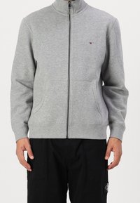 Grauer Sweatshirt zum Reißverschluss aus Baumwollmischung mit hohem Kragen, zwei Fronttaschen und einem kleinen Logoakzent auf der Brust. Glatte Textur.