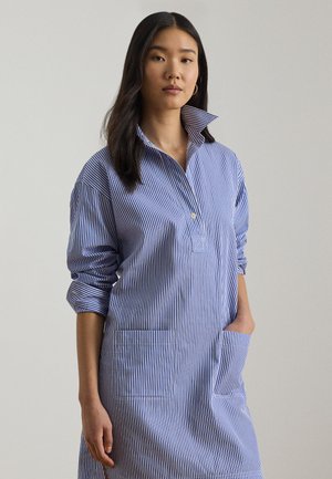 Mujer con cabello largo y oscuro que lleva un vestido a rayas verticales azul y blanco con las mangas remangadas, de pie con una mano en el bolsillo.