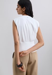 Haut blanc sans manches avec une taille cintrée et un détail de boutons dans le dos, associé à un pantalon beige à jambes larges et un bracelet noir épais.