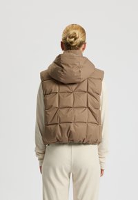 Braun gepolsterte Weste mit Kapuze, ausgestattet mit einer gesteppten Textur und einem hohen Kragen. Getragen über einem hellen, gerippten Pullover.