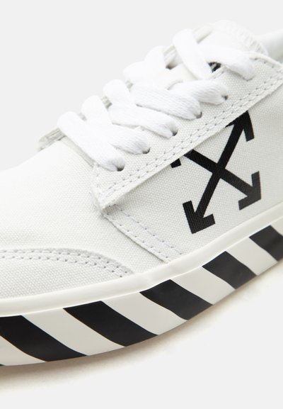 Λευκό αθλητικό παπούτσι με χοντρά λευκά κορδόνια, μαύρο λογότυπο βέλους σε σχήμα σταυρού στο πλάι και μαύρο-λευκή διαγώνια ριγέ άκρη της σόλας.