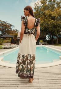 Robe maxi avec un design dos nu, dotée de manches volantes, de motifs de feuilles de palmier et d'une jupe à volants. La palette de couleurs comprend un beige clair et un vert foncé.