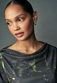 Rochie cu guler cowl din material închis, având un model cu pete multicolore de albastru și verde; modelul poartă cercei mici din argint în formă de cerc.