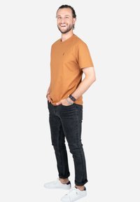 Bruine katoenen t-shirt, donkere jeans en witte sneakers. Model staat zijwaarts, glimlachend, met handen in de zakken. Minimalistisch ontwerp, casual pasvorm.