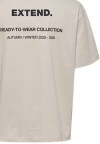 T-shirt in cotone beige con maniche corte, caratterizzato da testo nero: "EXTEND." e "COLLEZIONE PRONTA DA INDOSSARE AUTUNNO / INVERNO 2025 - 2026" sul retro.