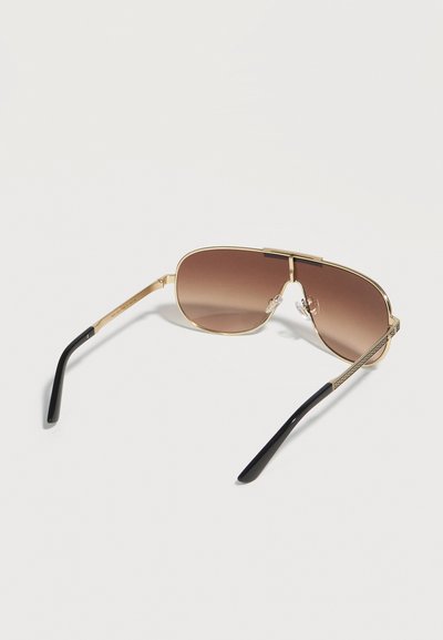 Lunettes de soleil aviateur avec monture dorée, verres dégradés marron et embouts d'oreille noirs reposent sur une surface blanche.