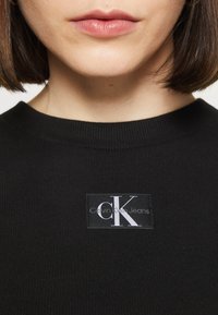 Černý žebrovaný svetr s čtvercovou nášivkou s textem "Calvin Klein Jeans" a s kontrastním lesklým nápisem "CK".