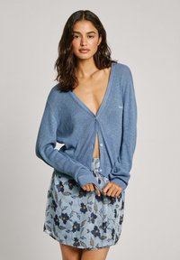 Zili rāvējslēdzēja džemperis ar dziļu V veida kakla izgriezumu, pogu priekšpusi un garām piedurknēm, uzlikts virs gaiši zilas ziedu raksta svārku.