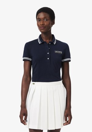 Mujer con polo azul marino con ribete blanco y falda plisada blanca, de pie contra un fondo liso.