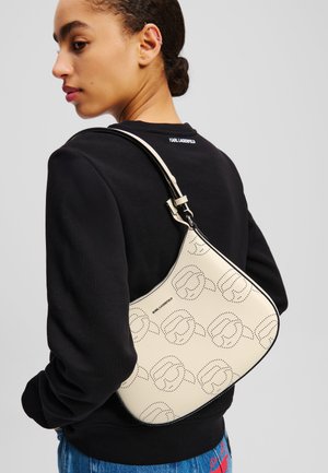 Borsa a tracolla crema con motivo logo perforato, forma curva e rifiniture nere; tracolla attaccata; indossata con una felpa nera.