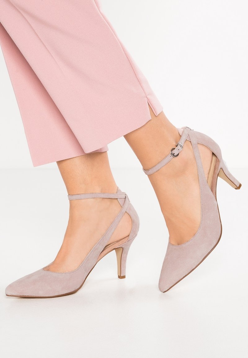 Des chaussures à talons hauts en suède gris clair avec un bout pointu, une bride de cheville et un design ouvert sur les côtés, sur fond blanc.