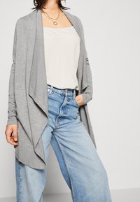 Gray cardigan with an open front and long sleeves, layered over a light beige top. Paired with high-waisted light blue jeans.