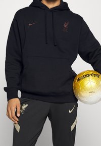 Sweat à capuche noir avec poche kangourou et cordon de serrage, arborant le logo Nike bordeaux et l'emblème de Liverpool FC. Pantalons gris avec des accents dorés, tenant un ballon de football doré.