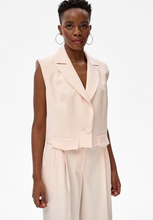 Kobieta o krótkich włosach, ubrana w bezrękawnikowy beżowy blezer i pasujące szerokie spodnie, stojąca na białym tle.