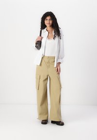 Pantaloni cargo beige con vestibilità oversize, dotati di molteplici tasche, abbinati a una blusa bianca e a una borsa nera. Il modello si trova contro uno sfondo bianco.