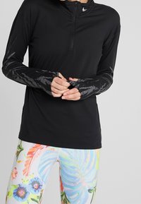 Osoba nosząca czarną, długą sportową bluzę z otworami na kciuki oraz kolorowe legginsy z dużymi, pastelowymi wzorami kwiatowymi.