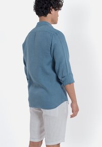 Camicia in lino azzurro chiaro con maniche lunghe arrotolate, pantaloni corti in lino bianco a contrasto, visti da dietro su uno sfondo neutro.