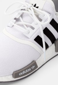 Zapatilla deportiva blanca con parte superior de malla texturizada, diseño de tres franjas negras, cordones blancos y suela gris con marca. Patrón de suela hexagonal visible.